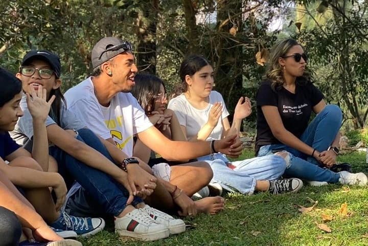 Foto de alguns jovens sorrindo sentados em circulo na grama em um dia ensolarado