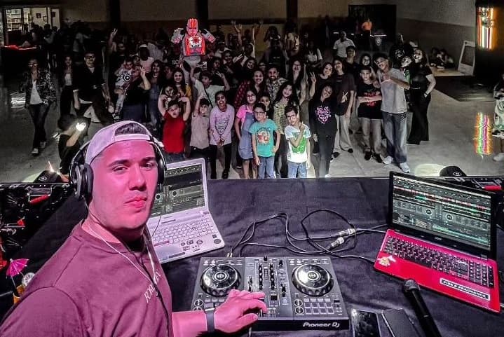 Foto de uma multidão de pessoas na frente de uma mesa de DJ com o DJ tirando uma selfie